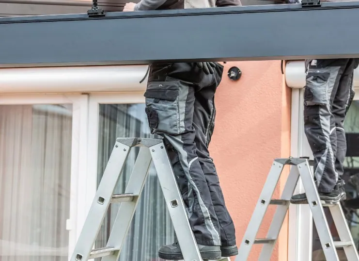 Dos técnicos subidos a escaleras montando el perfil de aluminio de una pérgola adosada a una vivienda